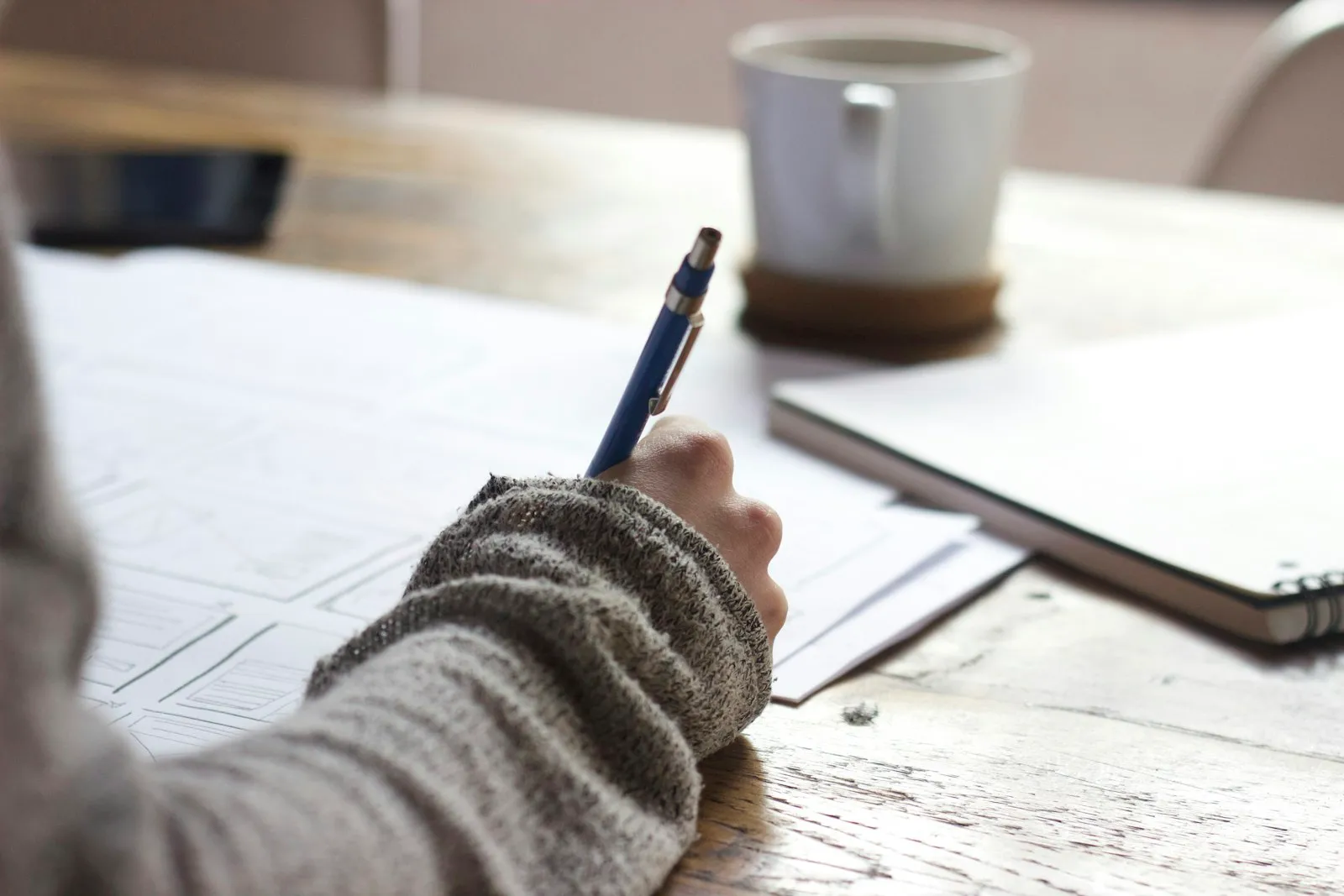 Hands writing notes at a wooden desk beside a coffee cup and laptop.